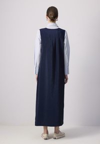 Vestido sin mangas de color azul marino con una textura suave, de largo extendido y con la parte trasera ligeramente fruncida, combinado con zapatillas beige.