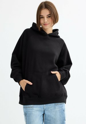 Sudadera negra de material suave, con bolsillo frontal, capucha y puños acanalados. Combinada con vaqueros azul claro.