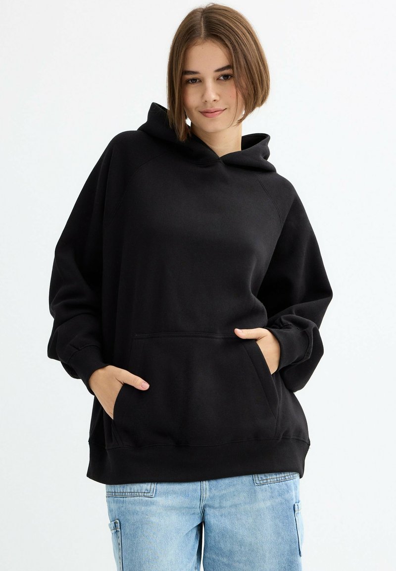 Sudadera negra de material suave, con bolsillo frontal, capucha y puños acanalados. Combinada con vaqueros azul claro.