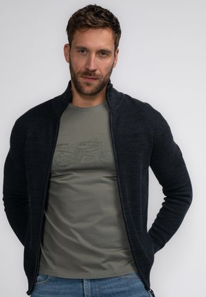 Donker marineblauwe gebreide cardigan met rits over een licht olijfgroen T-shirt met grafisch ontwerp, gecombineerd met lichtblauwe spijkerbroek.
