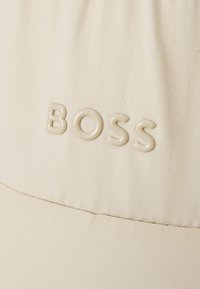 Beige kangri müts, mis on valmistatud kanvasest; esiküljel on reljeefne "BOSS" logo samas toonis. Äärel on nähtav õmbluse detail.