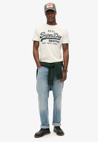 Camiseta blanca con texto de logo azul, vaqueros azul claro, suéter verde atado alrededor de la cintura y zapatos negros con suelas marrones. Gorra gris con logo rojo.