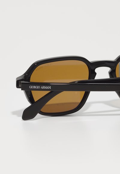 Ochelari de soare din plastic negru cu rame unghiulare și lentile de culoare maro. Bratul are inscripționat "Giorgio Armani" cu litere albe.
