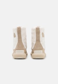 Beige ankelstøvler med en bløt, teksturert pelskropp, forsterket suedesåle, og gummisåle. Har en fremtredende "SOREL" logo på baksiden.