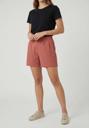 Femme portant un t-shirt noir à manches courtes, un short taille haute de couleur rouille et des baskets beiges, debout devant un fond clair uni.