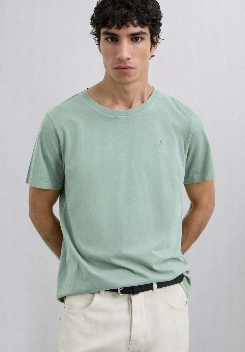 Joven con cabello rizado oscuro, vestido con una camiseta de cuello redondo de color verde claro y pantalones crema con un cinturón negro, de pie frente a un fondo liso.