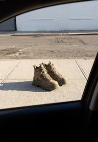 Paar hellbraune Schnürstiefel mit Tarnmuster auf einem Gehweg neben einer Straße, von innen im Auto gesehen.