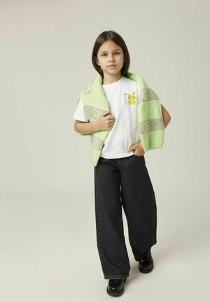 Camiseta blanca con un gráfico de Pikachu, suéter verde oversize caído sobre los hombros, jeans negros de pierna ancha, zapatos negros.