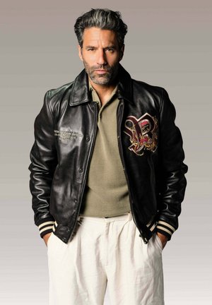 Homme aux cheveux poivre et sel portant une veste en cuir noire avec un blason brodé, un polo beige et un pantalon blanc, mains dans les poches, fond neutre.