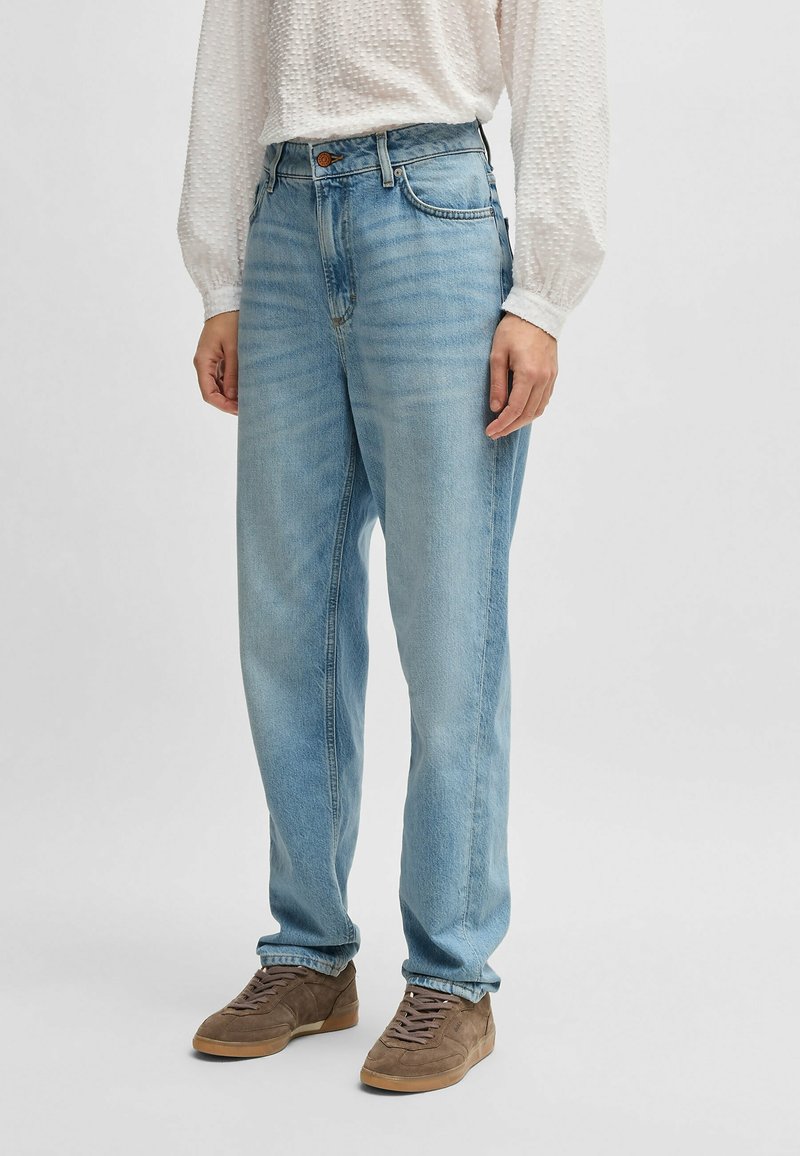 Osoba ubrana w jasnoniebieskie dżinsy o prostym kroju, białą, teksturowaną koszulę z długim rękawem oraz brązowe zamszowe sneakersy, stojąca na neutralnym tle.