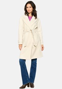 Beige wrap coat with a belt, double-breasted front, and large lapels, paired with blue straight-leg jeans and brown heeled ankle boots.