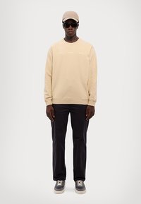 Béžová mikina s žebrovanými manžetami, černé kalhoty, šedé tenisky a béžová čepice. Sluneční brýle nasazené, stojí proti neutrálnímu pozadí.