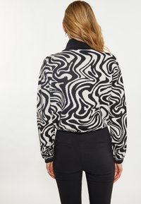 Schwarzer und weißer, wirbelmuster gestrickter Pullover mit hohem Kragen, langen Ärmeln und lockerer Passform, kombiniert mit schwarzen, figurbetonten Hosen.