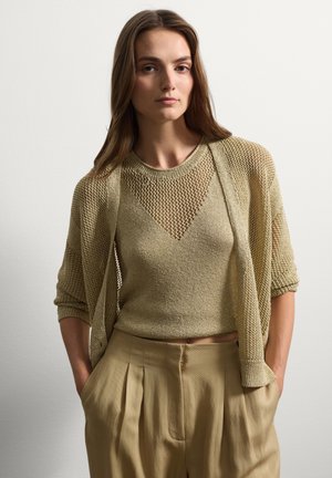 Jersey de punto en un tono oliva claro, con un diseño cropped y tejido abierto. Combinado con pantalones beige de talle alto y con pliegues.