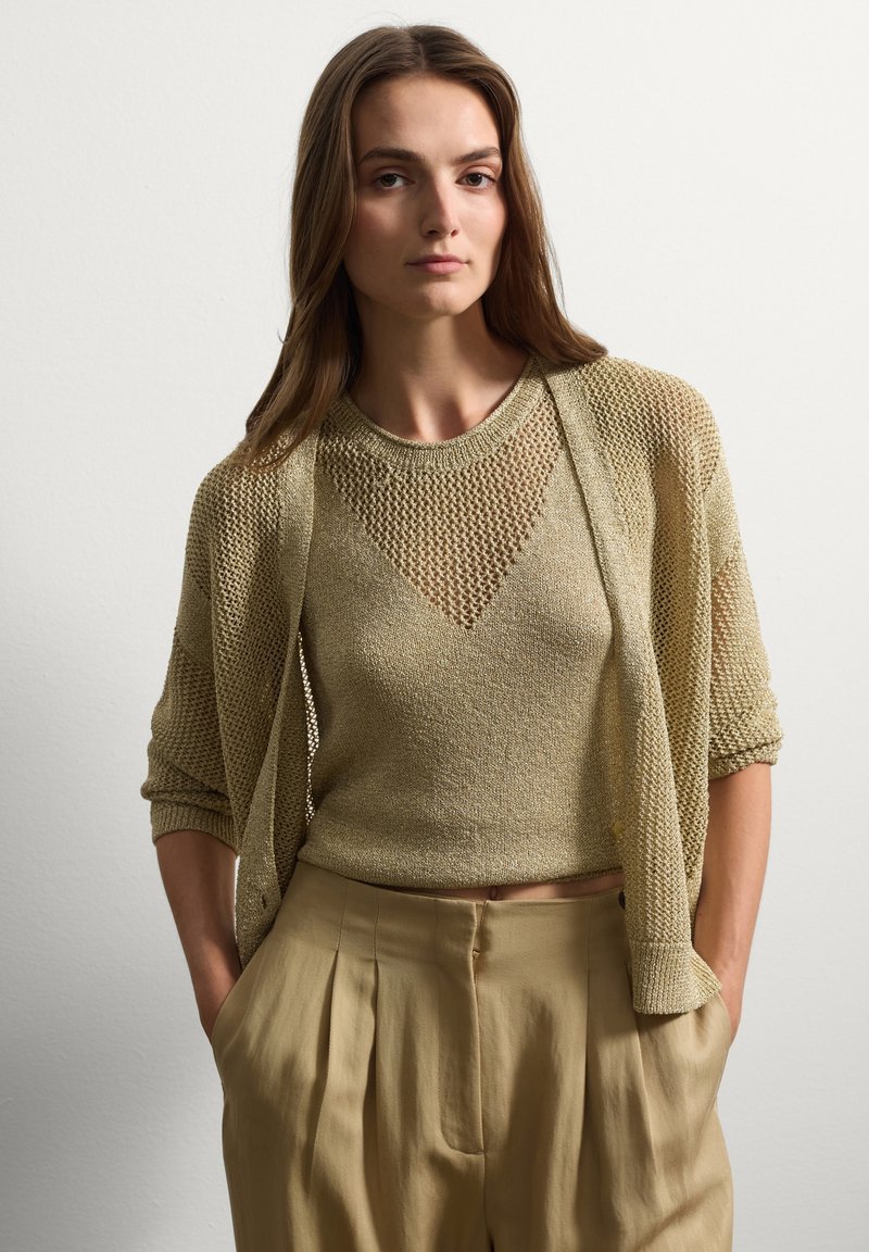 Maglione lavorato a maglia in oliva chiaro, con un design corto e trama aperta. Abbinato a pantaloni beige plissettati a vita alta.