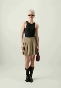 Top preto sem mangas, saia plissada cáqui, botas de cowboy pretas, bolsa castanha e óculos de sol; modelo de pé sobre um fundo claro.
