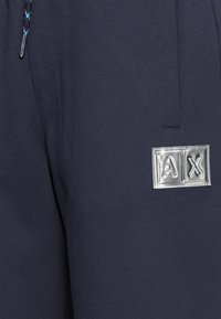 Marinblå joggingbyxor i mjukt tyg med en silverfärgad logotypatch "A|X" framtill, med en dragsko med ett mönstrat snöre.