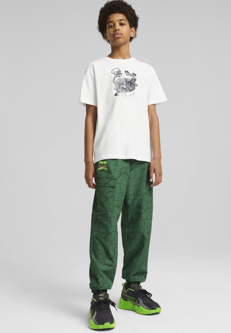 Hvid t-shirt med grafisk print, grønne sweatpants med spræk-mønster og sorte sneakers med grønne accenter. Afslappet, løs pasform.