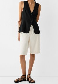 Svart ärmlös väst med djup V-ringning och tre knappstängningar, i kombination med lösa vita shorts och svarta sandaletter med remmar.