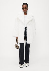 Biely kožuch z umelého zamatu so širokými klopami, v kombinácii s priliehavým bielym topom, čiernymi rozšírenými nohavicami a striebornými topánkami. Doplnkový malý vak s leopardím vzorom.