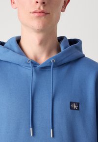 Modrá mikina s kapucňou z mäkkého materiálu, so znakový nášivkou a striebornými kovovými koncami šnúrky. Pohľad zblízka na výstrih.