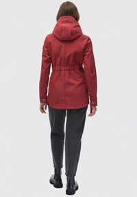 Rode waterdichte jas met een capuchon, een getailleerde taille, knopenmanchetten en een gladde textuur. Draagt een combinatie met donkergrijze broek en laarzen.