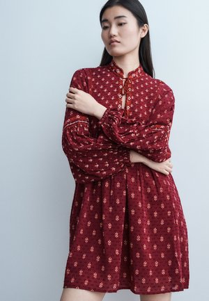 Mladá žena s dlhými tmavými vlasmi, oblečená v červenom šate so vzorom a dlhými nafúknutými rukávmi, stojaca pred jednoliatym svetlým pozadím.