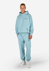 Svetlomodrá mikina s kapucňou a zodpovedajúce tepláky z mäkkej tkaniny. Obsahuje predné vrecko a čierny logo detail na oboch častiach. Materiál z bavlnenej zmesi.