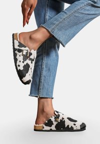 Sabots imprimés vache blancs et noirs avec semelle en liège, dos ouvert, détail de boucle en argent et intérieur en fourrure, associés à un jean léger bleu coupé.