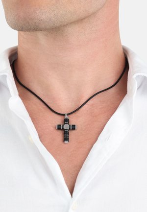 Pendente a croce nera realizzato con materiali testurizzati, appeso a una collana di corda nera, indossato sopra una camicia bianca con colletto.