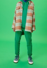 Person klädd i grön huvtröja och byxor med en beige och vit rutig yttertröja och grå sneakers som står mot grön bakgrund.