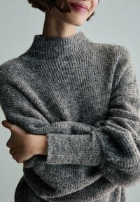 Maglione grigio lavorato a maglia con collo alto, caratterizzato da una texture a coste e polsini leggermente arrotolati. Il tessuto sembra morbido e accogliente.