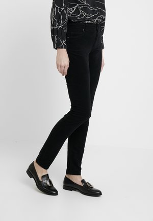 Pantalons skinny en velours côtelé noir avec une texture lisse, assortis à des mocassins noirs avec un accent métallique circulaire.
