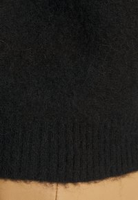 Svart strikket genserdetalj med en myk, teksturert overflate og ribbestrikket kant. Nærbilde viser fibre og sømmer mot en beige bakgrunn.