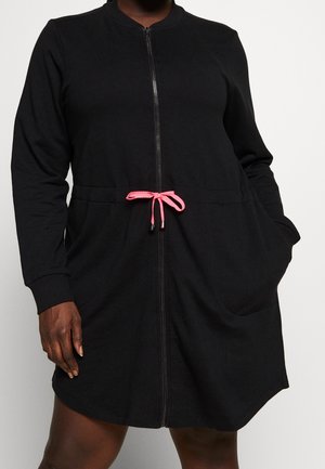 Person wearing a black zip-up dress with long sleeves, front pockets, and a pink drawstring tied at the waist.