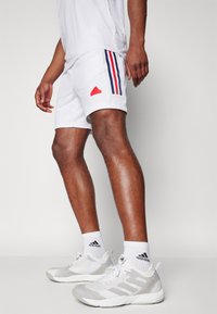 Shorts sportivi bianchi con strisce laterali rosse e blu, con logo Adidas rosso; abbinati a scarpe grigie e calze bianche.