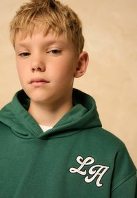 Sweat à capuche vert avec une poche frontale, comportant les lettres blanches brodées "LA" sur le côté gauche. Tissu doux, coupe décontractée et capuche avec cordons.