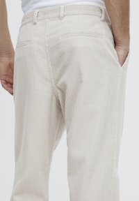 Light beige corduroy pants with vertical ribbing, two back pockets, and a standard waistband, showing a side view and hand in the pocket.