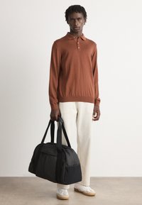 Person steht und hält eine große schwarze Duffel-Tasche, trägt ein rostfarbenes langärmliges Polohemd, cremefarbene Hosen und weiße Turnschuhe vor einem schlichten Hintergrund.