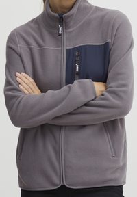 Giacca in pile grigia con una tasca sul petto con zip blu navy, colletto rialzato e tasche laterali con zip, caratterizzata da una texture morbida e un design aderente.