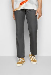 Pantalon droit gris à la texture lisse, associé à des baskets à lacets beiges avec des accents blancs. Les mains du mannequin sont visibles.