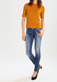 Top de manga corta de rayas en mostaza combinado con jeans ajustados azul desgastados y tacones negros. El atuendo presenta una silueta ajustada y un look casual.