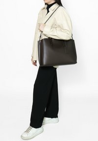 Sac fourre-tout en cuir marron au toucher lisse, de forme rectangulaire plate et avec de fines bretelles. Comprend une petite étiquette fixée sur le côté.