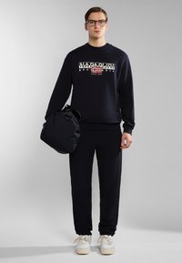 Felpa blu navy con logo, abbinata a joggers coordinati. Il modello tiene una borsa nera, indossando sneakers bianche con suola beige.