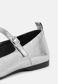 Silver metallic ballet flat with a textured finish, adjustable strap, and small black outsole. Compact and minimalist design.