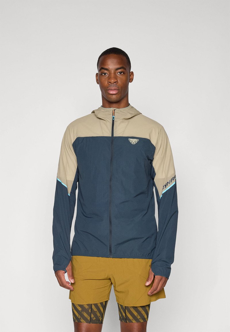 Windbreaker-Jacke mit einem beigefarbenen oberen Abschnitt und einem marineblauen unteren Teil, mit einem Reißverschluss, Kapuze und blauen Akzentdetails. Für den Einsatz im Freien konzipiert.