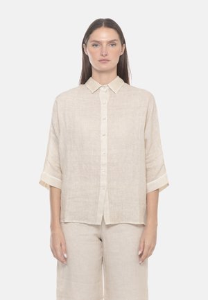 Donna con lunghi capelli castani che indossa una camicia di lino beige larga con bottoni e maniche arrotolate abbinata a pantaloni a gamba larga su sfondo bianco.