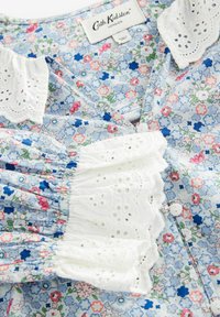Blusa floreale con uno sfondo azzurro chiaro decorato con fiori rosa e blu, rifinita con pizzo bianco ad occhiello, maniche a palloncino e chiusura a bottoni.