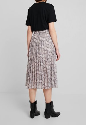 Femme portant un t-shirt noir, une jupe midi plissée à imprimé serpent et des bottines noires, debout devant un fond gris uni.