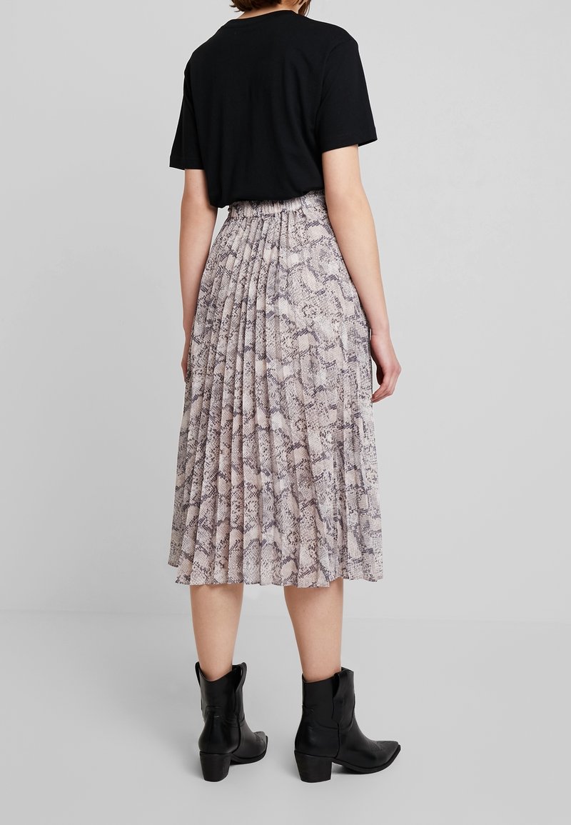 Femme portant un t-shirt noir, une jupe midi plissée à imprimé serpent et des bottines noires, debout devant un fond gris uni.